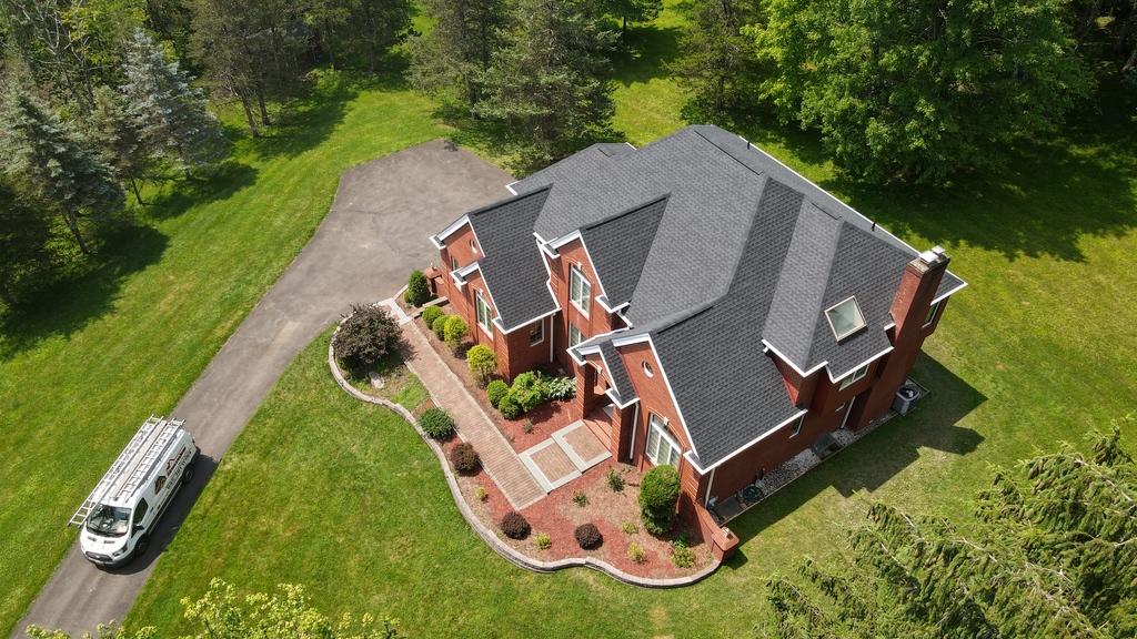 Aerial view of a large red brick house with black roof, surrounded by trees and grass, with a white utility van parked on the driveway.